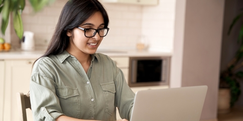 Smiling indian young adult woman wearing glasses typing on laptop computer working at home office sitting at table.