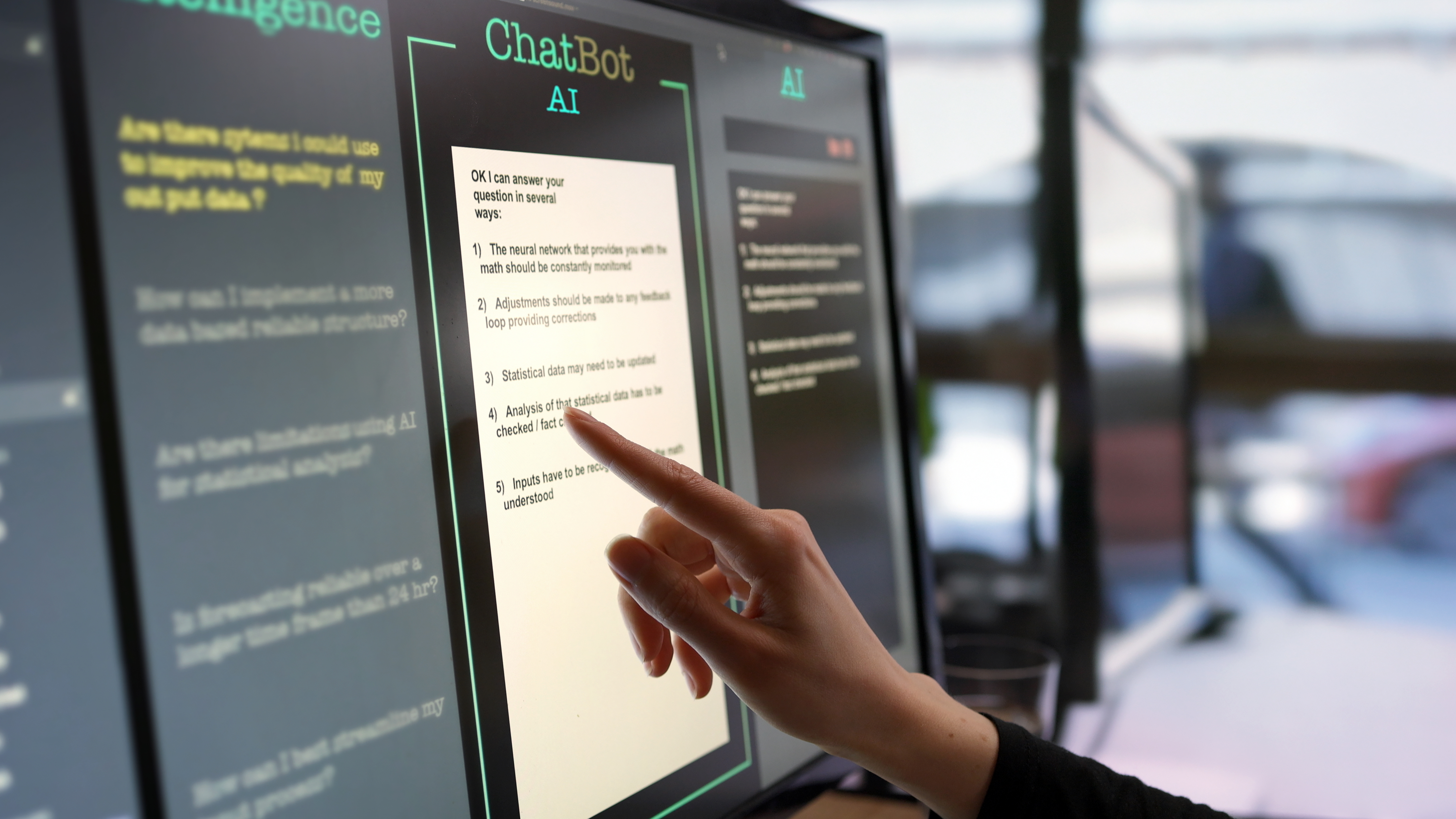 Close-up stock photograph showing a touchscreen monitor being used in an open plan office. A woman’s hand is asking an AI chatbot pre-typed questions & the Artificial Intelligence website is answering.