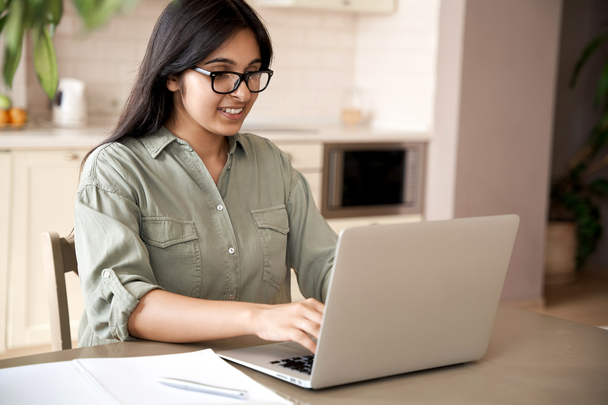 Smiling indian young adult woman wearing glasses typing on laptop computer working at home office sitting at table.